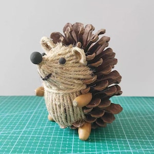 以下が含まれることがあります： 天然素材で作られた手作りのハリネズミの置物。体は紐で包まれ、黒いビーズの目と鼻が付いた頭があります。背中は茶色の松ぼっくりの鱗でできており、足はドングリでできています。置物は緑色のカッティングマットに飾られています。