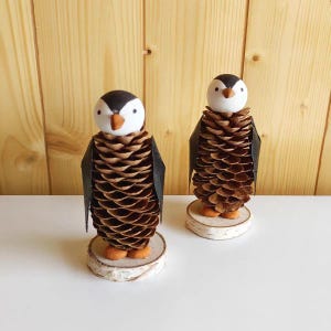 Könnte beinhalten: Zwei handgefertigte Pinguin-Figuren. Jeder Pinguin hat einen Körper aus Tannenzapfen, schwarze Flügel und einen weißen Kopf mit schwarzer Kappe und orangefarbenem Schnabel. Sie stehen auf kleinen Holzsockeln, ideal für die Dekoration.