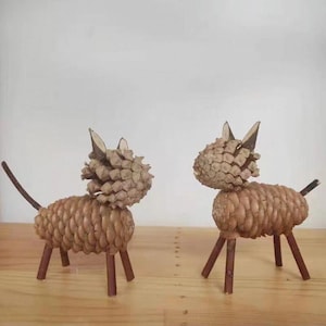 Pode incluir: Duas esculturas artesanais de animais, possivelmente gatos ou cães, feitas de pinhas e galhos. Os corpos são formados por pinhas, com pernas e caudas de galhos. As cabeças também são feitas de pinhas, com pequenas orelhas pontudas de galhos. As esculturas são marrons e estão sobre uma superfície de madeira.