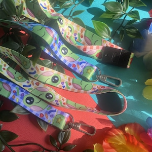 May include: Several colorful lanyards with a cartoon frog design. The lanyards feature a green frog face with black eyes and are decorated with confetti and stars. Each lanyard has a silver-colored clasp and a black plastic clip.