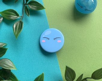 May include: A blue and pink button with a cartoon face. The face has closed eyes and a surprised expression. The button is on a green and blue background.