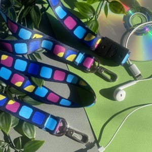 May include: Three colorful lanyards with a repeating pattern of blue, pink, and yellow squares on a navy background. Each lanyard has a silver-colored clasp. A black connector, a white earbud, and a CD are also visible.