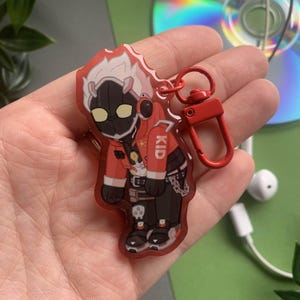 May include: A red and black anime character keychain with white hair and a red jacket. The character is wearing a black helmet with a yellow visor and has the word "KID" written on their jacket. The keychain is attached to a red metal clasp.