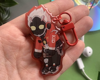 May include: A red and black anime character keychain with white hair and a red jacket. The character is wearing a black helmet with a yellow visor and has the word "KID" written on their jacket. The keychain is attached to a red metal clasp.
