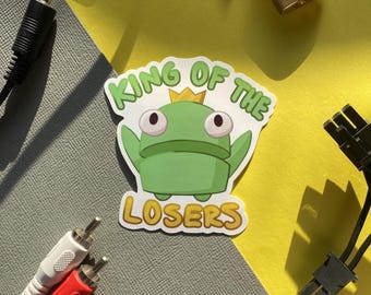Könnte beinhalten: Ein grüner Frosch mit einer goldenen Krone auf einem weißen Aufkleber mit dem Text "King of the Losers".