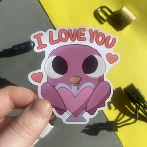 May include: A pink sticker with the words "I LOVE YOU" in red lettering. The sticker features a cartoon bird holding a heart. Two small hearts are on either side of the bird. The background is yellow and gray.