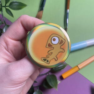 May include: A round button with a cartoon fish design. The button has a yellow and orange background with a fish illustration in the center. The button is held by a hand, with art supplies in the background.