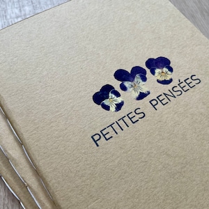 Op de afbeelding: Een bruin notitieboekje met de tekst "PETITES PENSÉES" gedrukt op de omslag. Drie gedroogde paarse en witte viooltjes zijn in een rij boven de tekst gerangschikt.
