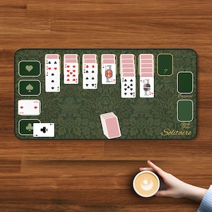 May include: A green Solitaire playing mat with a floral pattern, featuring playing card layouts and the text "Stock Pile Solitaire." A hand holds a cup of coffee in the foreground. The mat displays playing cards.