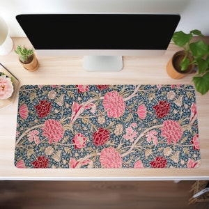May include: A rectangular desk mat with a floral design, showcasing large pink and red flowers against a dark blue backdrop. Beige vines and small white flowers complete the pattern. The mat sits on a light wood desk, with a computer monitor and plants in view.