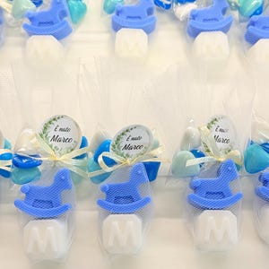 Puede incluir: Recuerdos de baby shower con decoraciones de caballito balancín azul, caramelos blancos con la letra "M" y pequeños caramelos azules y blancos. Cada recuerdo está envuelto en celofán transparente y atado con una cinta. Los recuerdos tienen una etiqueta que dice "È nato Marco".