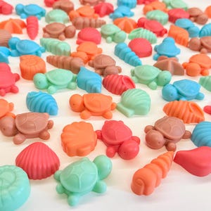 May include: A collection of small, solid-coloured sea-themed items shaped like turtles, starfish, and various seashells. The pieces are rendered in a vibrant palette of coral, turquoise, mint green, and soft pink, scattered across a plain surface.