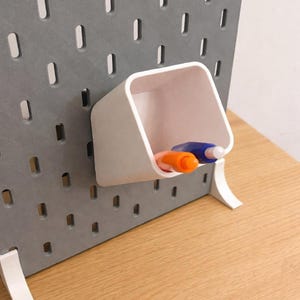 Puede incluir: Un panel perforado gris con un portalápices blanco y dos bolígrafos. El portalápices es cuadrado e inclinado, con un bolígrafo naranja y otro azul en su interior. El panel perforado tiene varios agujeros rectangulares y está sostenido por patas blancas.