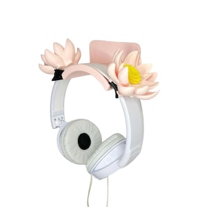 May include: White headphones with light pink accents and decorative pink and yellow flower accents. The headphones are hanging on a light pink holder, creating a floral and modern aesthetic.