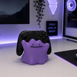 Pode incluir: Um suporte para comando roxo em forma de gota, com um rosto simples composto por dois pontos pretos para os olhos e uma linha horizontal para a boca. O suporte segura um comando de jogo sem fios preto com botões e joysticks visíveis.