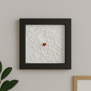 Könnte beinhalten: Ein gerahmter, weißer 3D-Stadtplan mit schwarzem Rahmen. Ein kleiner, roter Herzstift ist auf der Karte platziert. Die Karte zeigt ein Stadtlayout mit Straßen und Gebäuden. Der Rahmen ist quadratisch und an einer hellgrauen Wand befestigt.
