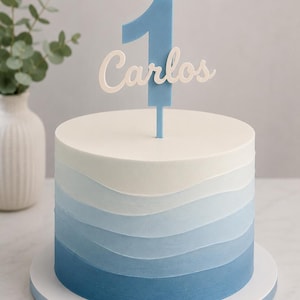 Pode incluir: Um bolo redondo com camadas de cobertura azul degradê horizontal, passando do branco no topo ao azul escuro na base. Um topo de bolo de acrílico azul em forma de número um com o nome Carlos em letra cursiva branca está inserido.