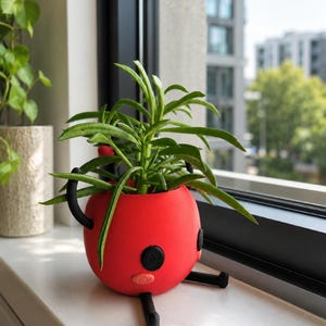 May include: A red, spherical plant pot with black detailing and legs, containing a green plant with long, thin leaves. The pot is on a white windowsill, with a blurred cityscape visible through a window in the background.