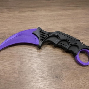May include: A curved karambit knife with a purple blade and ring, and a black handle. The knife is designed with a finger ring for secure grip. The blade has a sharp, curved shape.
