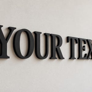 Op de afbeelding: Zwarte driedimensionale letters die de woorden YOUR TEXT vormen in een schreeflettertype. De letters zijn afzonderlijk gemonteerd, wat een verhoogd effect creëert met strakke randen en een matte afwerking voor een heldere typografische weergave.