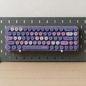 May include: A purple and pink MOFII keyboard with round keys is mounted on a gray pegboard. The keyboard has white, pink, and purple keys. The pegboard is supported by white legs and sits on a wooden surface.