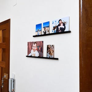 May include: Two horizontal black wall-mounted floating shelves displaying five rectangular photo prints. The top shelf holds three vertical prints, while the bottom shelf holds two prints, one horizontal and one vertical, featuring various family portraits.