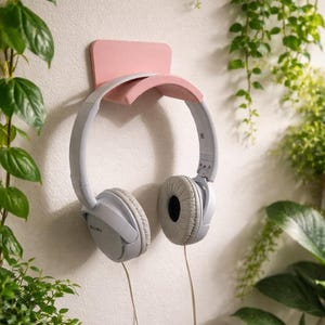 Pode incluir: Um par de fones de ouvido Sony cinza claro pendurados em um suporte de parede rosa. O suporte está montado em uma parede branca, rodeado por plantas verdes. O cabo dos fones de ouvido é visível.