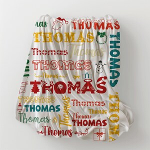 May include: A white blanket with the name "Thomas" in various fonts and colors, including red, yellow, and teal. Christmas-themed graphics such as a Santa hat, snowman, and gift boxes are also featured.