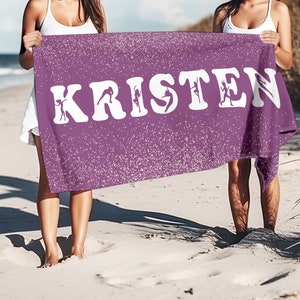May include: Two people are holding up a purple beach towel with white glitter and the name "KRISTEN" written in white letters. The letters are outlined with silhouettes of dancers.