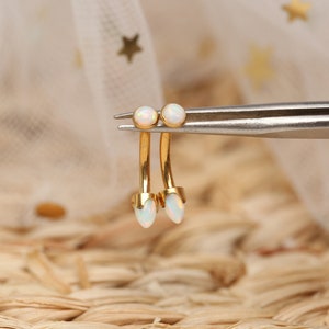 May include: Two gold body jewellery pieces with white opal stones. The jewellery is held by tweezers.