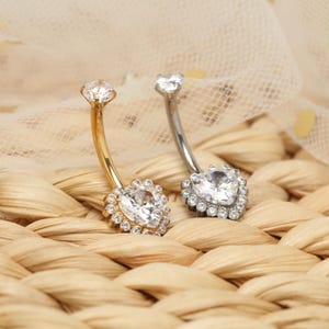 May include: Two belly button rings. One is gold-coloured, the other silver-coloured. Both have a curved barbell design. The lower charm is a heart-shaped clear stone surrounded by smaller clear stones. The top charm is a single clear stone.