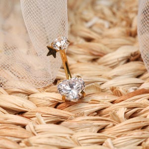 May include: Gold belly button ring with a clear heart-shaped gem and a smaller clear gem on a star-shaped charm.