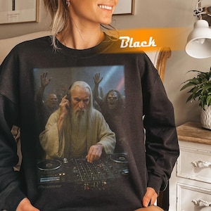 Pode incluir: Camisola preta com um gráfico engraçado de um DJ com uma longa barba branca e uma multidão de zumbis ao fundo. O DJ está usando fones de ouvido e operando um mixer Pioneer DJ. A palavra "Black" está impressa em laranja.