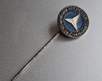 May include: A vintage Mercedes-Benz lapel pin. The pin features a circular emblem with a blue background and a silver star. The emblem is surrounded by a silver wreath and the words "MERCEDES BENZ". The pin has a long, silver needle.
