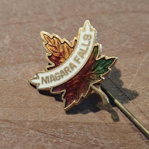 Peut inclure: Une épingle dorée en forme de feuille d'érable avec l'inscription "Niagara Falls" sur une bannière blanche. La feuille présente des détails en émail rouge, orange et vert. L'épingle a une longue et fine aiguille.