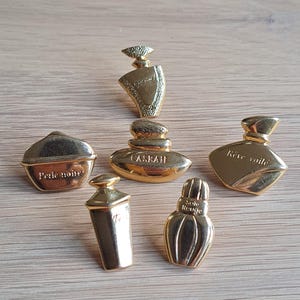 Pode incluir: Uma coleção de seis broches dourados em forma de frasco de perfume. Cada broche tem um design e forma únicos, com texto em alguns deles. Os broches estão dispostos sobre uma superfície de madeira castanha clara.