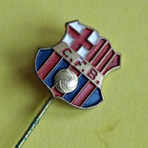Puede incluir: Pin vintage con el escudo del FC Barcelona. El emblema en forma de escudo tiene rayas rojas y azules, una cruz blanca y un balón de fútbol dorado. Las letras "C.F.B." son visibles. El pin tiene un vástago verde.