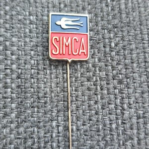 May include: A vintage SIMCA pin with a rectangular shape. The top half is blue with a white bird emblem, and the bottom half is red with the word "SIMCA" in white. The pin has a long, thin, gold-colored needle.