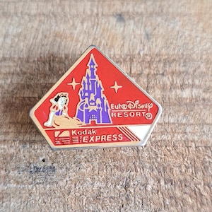 May include: A red, white, and gold enamel pin featuring a purple castle, a woman in a blue and white dress, and the text "Euro Disney Resort" and "Kodak Express".