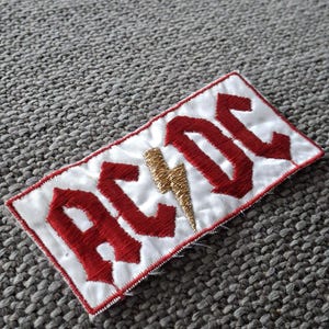 Peut inclure: &Eacute;cusson brod&eacute; avec le nom du groupe "AC/DC" en lettres rouges sur fond blanc. Un &eacute;clair dor&eacute; est plac&eacute; entre les lettres. Le patch a une bordure cousue rouge.