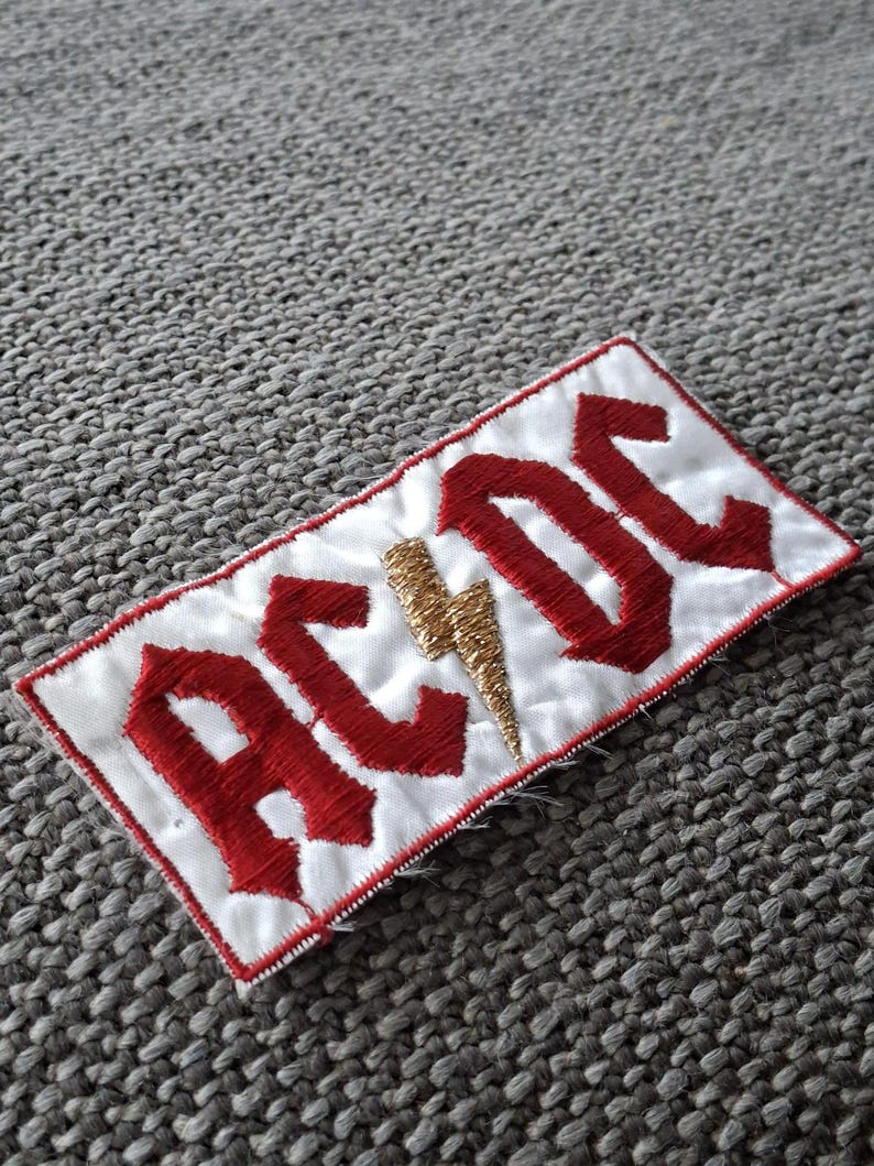 Peut inclure: &Eacute;cusson brod&eacute; avec le nom du groupe "AC/DC" en lettres rouges sur fond blanc. Un &eacute;clair dor&eacute; est plac&eacute; entre les lettres. Le patch a une bordure cousue rouge.
