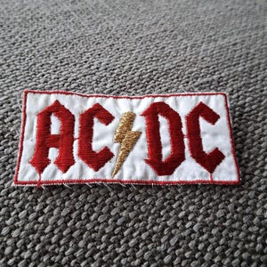 Peut inclure: &Eacute;cusson brod&eacute; avec le texte "AC/DC" en rouge, lettres gothiques sur fond blanc. Un &eacute;clair dor&eacute; est plac&eacute; entre les lettres. L'&eacute;cusson a une bordure rouge et est de forme rectangulaire.