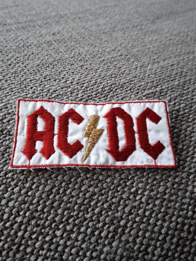 Peut inclure: &Eacute;cusson brod&eacute; avec le texte "AC/DC" en rouge, lettres gothiques sur fond blanc. Un &eacute;clair dor&eacute; est plac&eacute; entre les lettres. L'&eacute;cusson a une bordure rouge et est de forme rectangulaire.