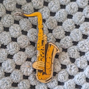 Pode incluir: Um alfinete de saxofone amarelo com um design detalhado, apresentando uma mão segurando o instrumento. O alfinete está sobre um fundo azul claro texturizado.