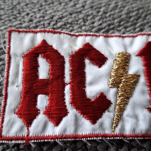 Peut inclure: &Eacute;cusson brod&eacute; avec les lettres "AC/DC" en rouge, police gothique, et un &eacute;clair dor&eacute;. L'&eacute;cusson est sur un fond blanc avec une bordure rouge.