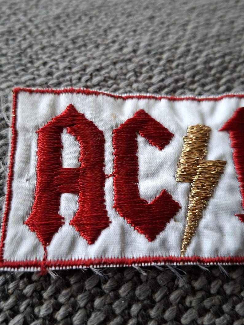 Peut inclure: &Eacute;cusson brod&eacute; avec les lettres "AC/DC" en rouge, police gothique, et un &eacute;clair dor&eacute;. L'&eacute;cusson est sur un fond blanc avec une bordure rouge.