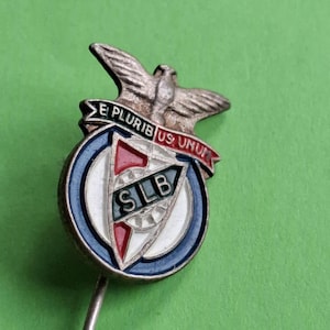 May include: A silver-coloured pin badge with a circular design in blue, white, and red. The badge features the letters "SILB", an eagle, and a banner with the inscription "E PLURIBUS UNUM".