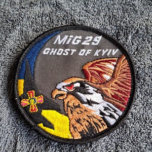 Puede incluir: Parche bordado circular con un diseño de halcón y el texto "MiG 29 Ghost of Kyiv". El parche tiene un borde negro y presenta los colores amarillo, azul, rojo y marrón. El halcón está representado con plumas detalladas.