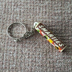 Könnte beinhalten: Ein zylindrischer Schlüsselanhänger mit einer braunen Verpackung, auf der der weiße Schriftzug SMARTIES aufgedruckt ist, verziert mit bunten Bonbon-Illustrationen. Der Schlüsselanhänger enthält einen silberfarbenen Metallring an einer kurzen Kette.
