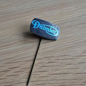Può includere: Spilla Daimler vintage con una testa rettangolare nera e la scritta "Daimler" in blu. La spilla ha un ago lungo e sottile color oro. La spilla è appoggiata su una superficie di legno.